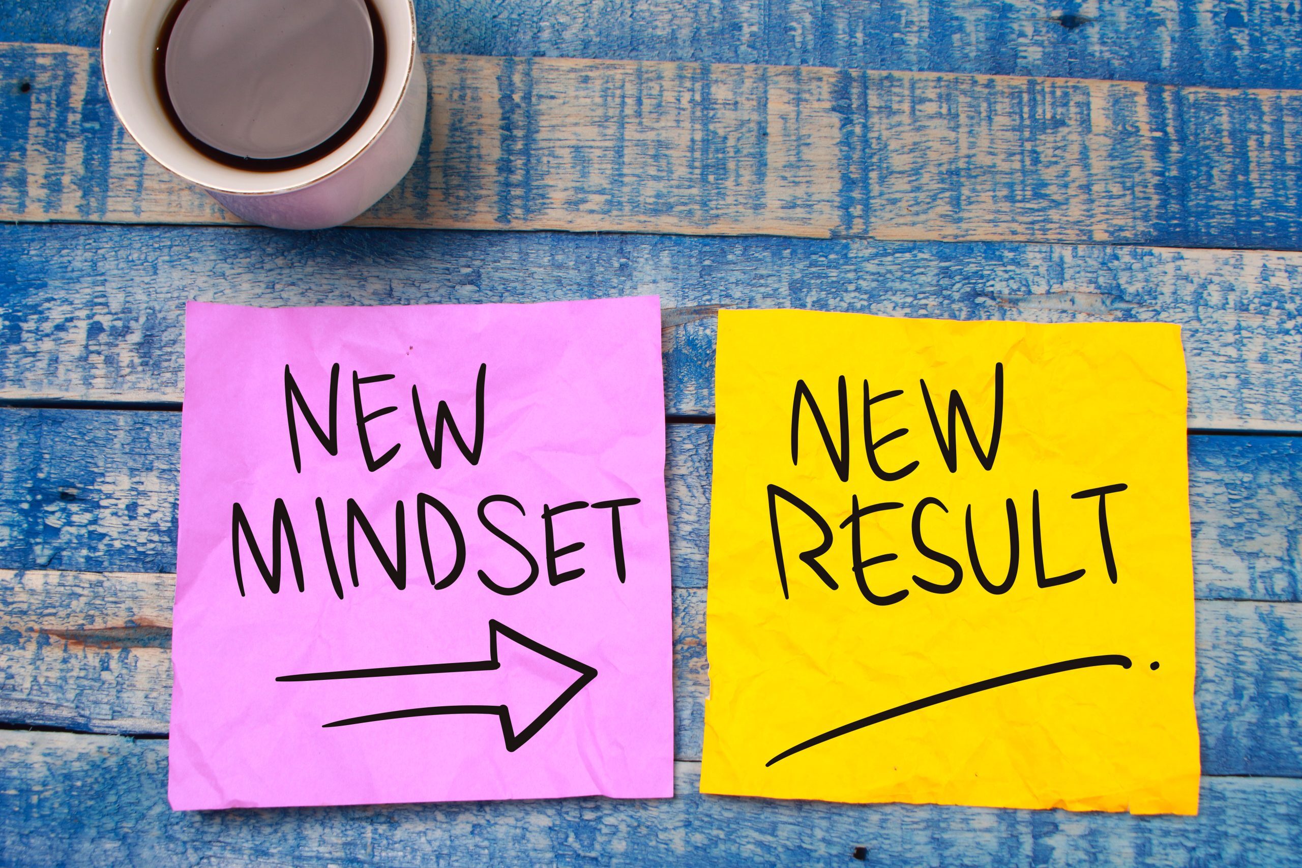 two post-it notes on a blue wood table, first post-it says "new mindset" with arrow leading to "new result" on second post-it